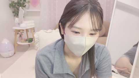 Video of an Asian woman with long black hair and light skin wearing a blue shirt and white mask, sitting on a bed in a pastel-colored room with a plant, mirror, and decorative items.