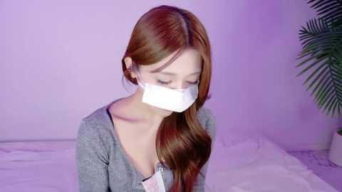 Video of a young Asian woman with long brown hair, wearing a white surgical mask, grey cardigan, and sitting on a bed in a softly lit room with purple walls.