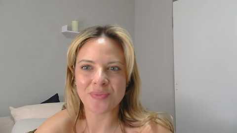 Video of a smiling Caucasian woman with blonde hair, fair skin, and blue eyes, wearing a white top. She's in a minimalistic, light-colored bedroom with a white door and a shelf with candles in the background.