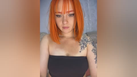 A video of a fair-skinned woman with vibrant orange hair and heavy makeup, wearing a strapless black top, revealing a detailed floral tattoo on her left shoulder.