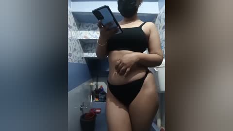 A video of a young woman with medium skin tone, wearing black lingerie, standing in a bathroom with blue and white wallpaper, taking a selfie in the mirror.