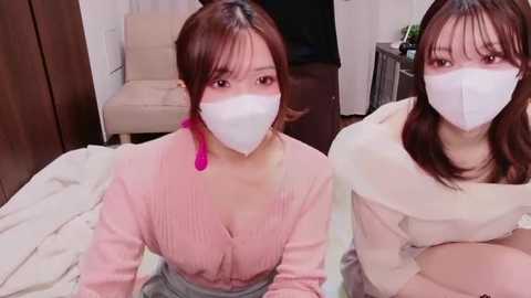 Video of two East Asian women with fair skin, wearing white face masks, pink ribbed sweaters, and grey skirts, sitting on a bed in a modern bedroom.