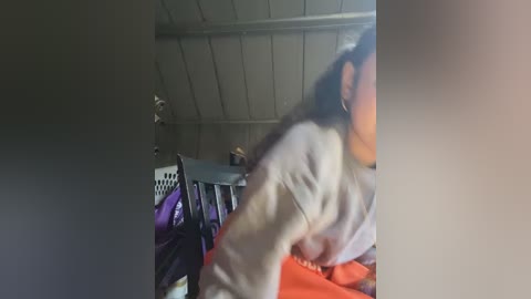 Video of a woman in a white sweater and orange pants, blurred, leaning over a laundry basket filled with clothes in a dimly lit attic room with a slanted ceiling and wooden beams.