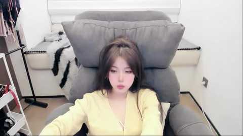 A video of an Asian woman with straight black hair, wearing a yellow cardigan, reclining in a grey armchair. Behind her, a white window, a black-and-white blanket, and a clothing rack with items are visible.