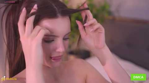Video of a young, light-skinned woman with long brown hair, wearing minimal makeup, combing her hair with her hands. She is topless. Background shows blurred greenery and a soft-focus headboard.