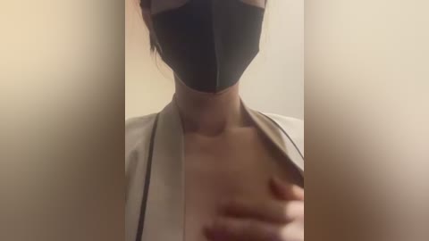 Media: A video of a person with light skin, wearing a black face mask, beige top, and visible collarbone. The background is blurred, with a neutral beige tone.