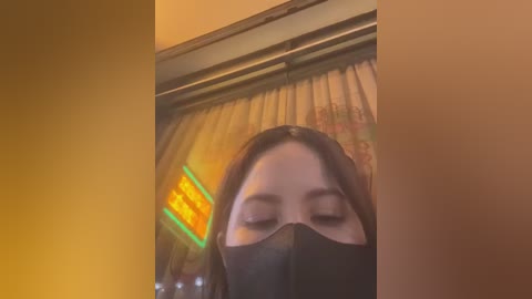 Media: A video of a woman wearing a black face mask, with her face partially obscured, standing in front of a dimly lit, beige curtain. A neon sign with yellow text and green accents is visible in the background.