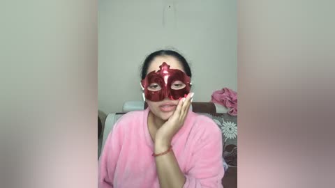 A video of a woman with medium skin tone, wearing a red masquerade mask, pink bathrobe, and red nails, sitting on a bed in a dimly lit room.