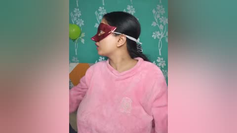 Video of an Asian woman with long black hair, wearing a pink sweater and red cardboard glasses, playing with a green ball against a turquoise wall with white floral patterns.