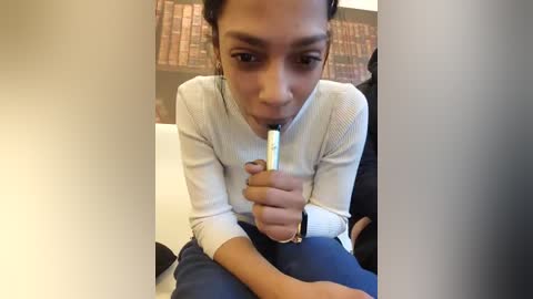 Video of a young woman with light brown skin and dark hair in a ponytail, holding a pen to her mouth, sitting on a white couch. Background features a bookshelf filled with books.
