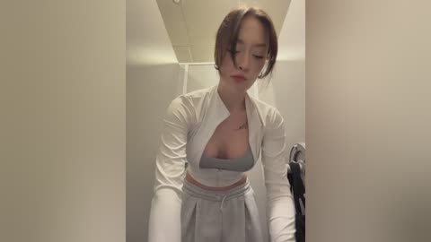 Video of a young woman with fair skin and brown hair styled in loose waves, wearing a white cropped top and high-waisted pants, standing in a narrow hallway with beige walls and a white ceiling.