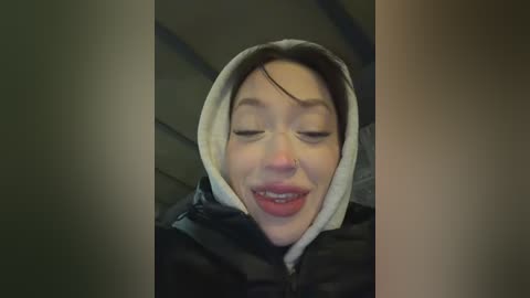 Video of an Asian woman with light skin, dark hair, and a nose ring, smiling with eyes closed, wearing a black and white puffer jacket. Background is dark and blurry.