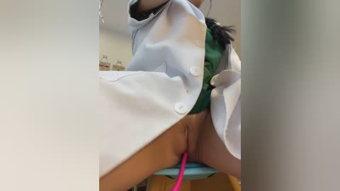 Video of a woman with light skin, wearing a white lab coat, green bra, and pink vibrator, sitting on a chair in a medical office setting.