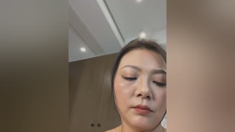 Video of a young East Asian woman with fair skin and straight, dark hair, wearing light makeup, looking downwards. Background shows a modern, beige-toned room with recessed ceiling lights and a wooden wardrobe.