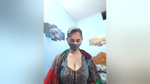 Video of a woman with dark skin, wearing a gray face mask, red shawl, and a multicolored patterned dress. Background features light blue walls with colorful, abstract art and a wooden headboard.