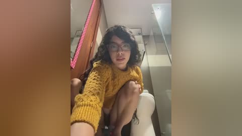 A video of a young person with long, curly black hair, wearing glasses and a mustard-yellow sweater, squatting in a modern bathroom with a white toilet and beige walls.