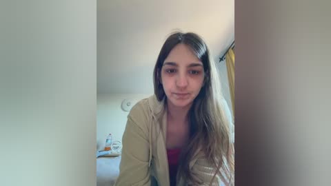 A video of a young woman with long brown hair, fair skin, and a slender build, wearing a beige robe and pink top, standing in a dimly lit bathroom with a shower curtain partially visible in the background.