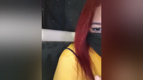 A video of a woman with long, straight red hair, wearing a black mask, and a yellow shirt, seen from the side, with a blurred background.