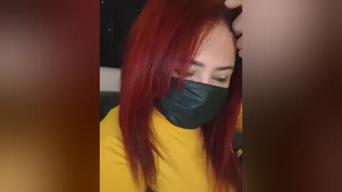 A video captures a woman with long red hair, wearing a yellow top and a black face mask, seated in a dimly lit room.