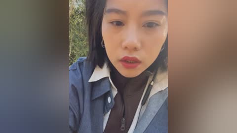 Video of a young Asian woman with medium skin tone, wearing a denim jacket over a black shirt, with shoulder-length dark hair, standing outdoors against a blurred green background.