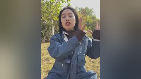 Video of an Asian woman with long black hair, fair skin, and red lipstick, wearing a dark blue coat, making a peace sign outdoors in a grassy area with trees.