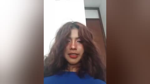 Video of a young Asian woman with wavy, shoulder-length brown hair, wearing a blue top, smiling. Background includes a white wall and a wooden door.