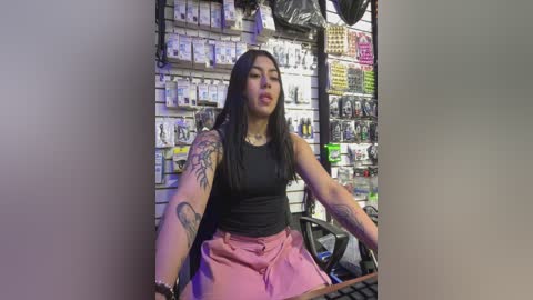 Video of a tattooed Latina woman with long black hair, wearing a black tank top and pink shorts, sitting behind a counter in a brightly lit store filled with electronic gadgets.
