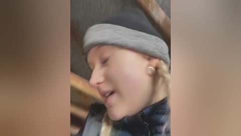 Video of a young blonde girl in a gray beanie, smiling, wearing a blue plaid shirt, with a blurred background of a wooden structure.