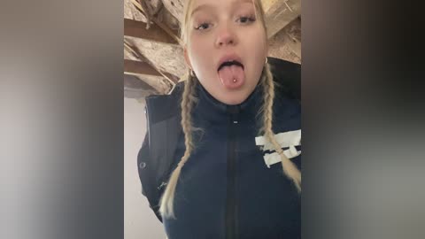 A video shows a young woman with blonde pigtails sticking out her tongue, wearing a navy jacket with a white emblem, in a dimly lit attic with exposed wooden beams.