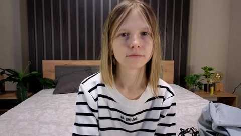 Video of a young blonde girl with straight hair, wearing a black-and-white striped shirt, sitting on a bed with a wooden headboard, dark curtains, and potted plants on either side.