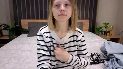 Video of a young Caucasian girl with straight, shoulder-length blonde hair, wearing a white and black striped sweater. She sits on a bed with a grey quilt, holding a black cord. The room has a modern, minimalist vibe with a wooden headboard and grey curtains.