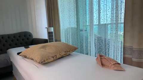 Video of a minimalist, modern bedroom with a single bed featuring a beige pillow and a folded pink blanket. The background showcases sheer curtains and a gray upholstered chair.