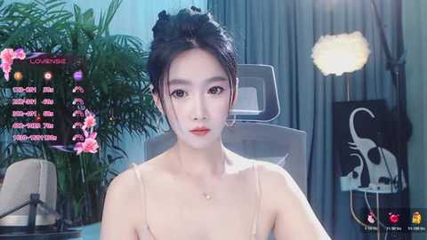 Video of a young Asian woman with fair skin and black hair in an updo, wearing a beige camisole, sitting in a modern room with a green plant, a lamp, and a large monitor displaying a pink floral background.