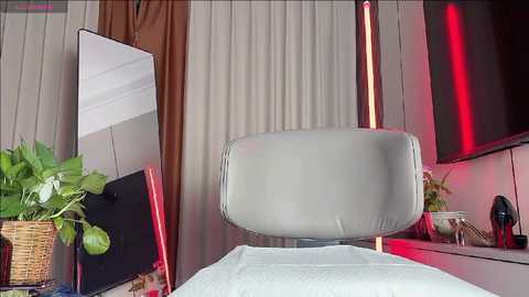 Media: Video of a modern bedroom with a sleek, white leather chair, a potted plant, a tall vertical mirror, a flat-screen TV, and red lighting accents on the wall.