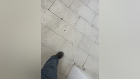 A video of a person's legs in blue jeans and black shoes walking on a tiled floor with light gray marble tiles, showing a diagonal pattern and slight wear. The background is a plain white wall.