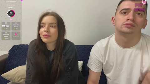 Video of a young Caucasian woman with long brown hair, wearing a black top, and a young man with short dark hair, wearing a white t-shirt, sitting on a dark blue couch, indoor setting.