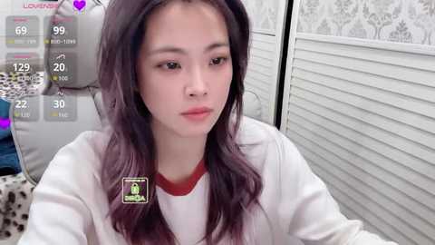 A video of a young East Asian woman with long, wavy dark hair and fair skin, wearing a white hoodie, seated indoors. The background features white louvered doors and a leopard print cushion.