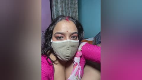 Video of a woman with medium skin tone, wearing a pink long-sleeve top, gray face mask, and red dot on forehead, sitting indoors with blurred background.
