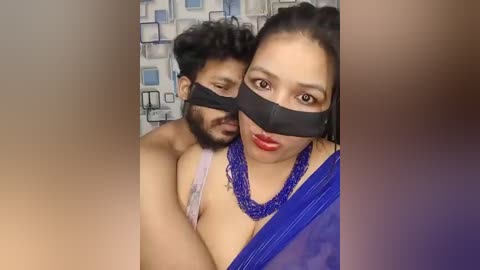 Video of a South Asian man and woman wearing black masks, the woman in a blue sari, the man shirtless, in a room with patterned wallpaper.