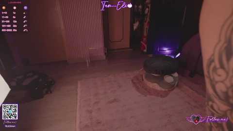 A dimly-lit video of a living room with a small, dark-haired woman in a pink robe kneeling on a rug, a tattooed arm visible.