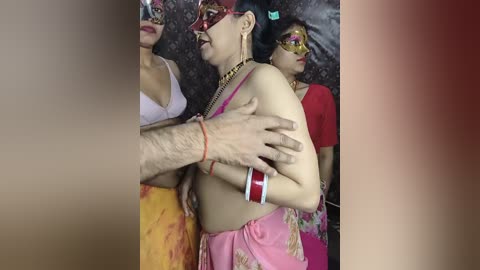 Video of a group of women in colorful saris and masks, with a man's arm around a woman's chest.