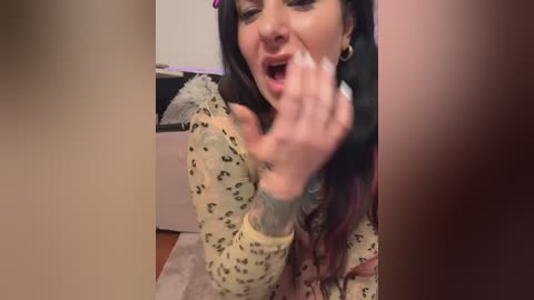 Video of a woman with long, dark hair and a leopard print top, mouth open, hand to her face, in a blurred, indoor setting.