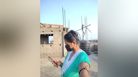 Video of a woman with dark hair in a ponytail, wearing a green dress, face mask, and bracelet, standing outdoors in front of a rustic brick wall and a windmill under a clear blue sky.