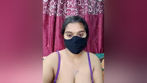 Video of a South Asian woman with medium skin tone, dark hair, wearing a black mask, purple bra, and a necklace, standing against a burgundy curtain with white floral pattern.