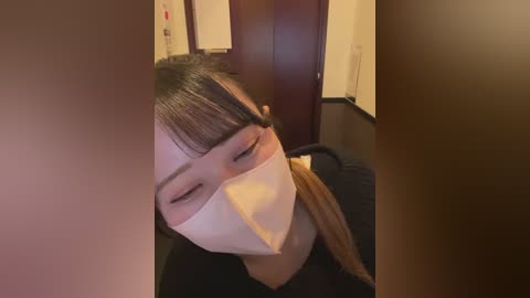 Video of an Asian woman with short black hair and bangs, wearing a white face mask and a black shirt, looking down, indoors.