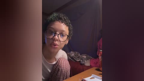 Media: A video of a curly-haired, fair-skinned woman with glasses, wearing a striped shirt, peering through a door frame, with a messy room in the background.