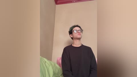 Video of a young person with short green hair, glasses, wearing a black sweater, standing in a beige-walled room with a pink and green bed.