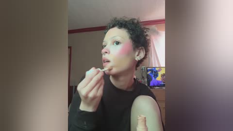 Video of a woman with curly hair, wearing a black top, applying lipstick in a bedroom with a TV showing a cartoon in the background.