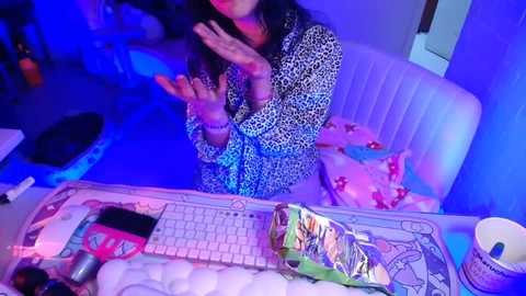 A video of a woman with long black hair in a leopard-print shirt, clapping, sitting at a desk with a keyboard, colorful bag, and cup. Room bathed in blue light.