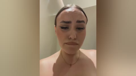 Video of a young woman with light skin, dark eyebrows, and long eyelashes, wearing a gold necklace, standing in a beige room with a lamp and wall.